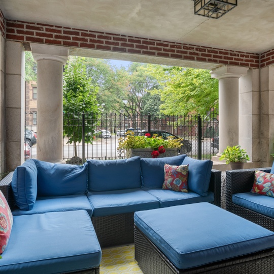 Covered patio with blue cushioned wicker seating and a view of a leafy fenced garden.